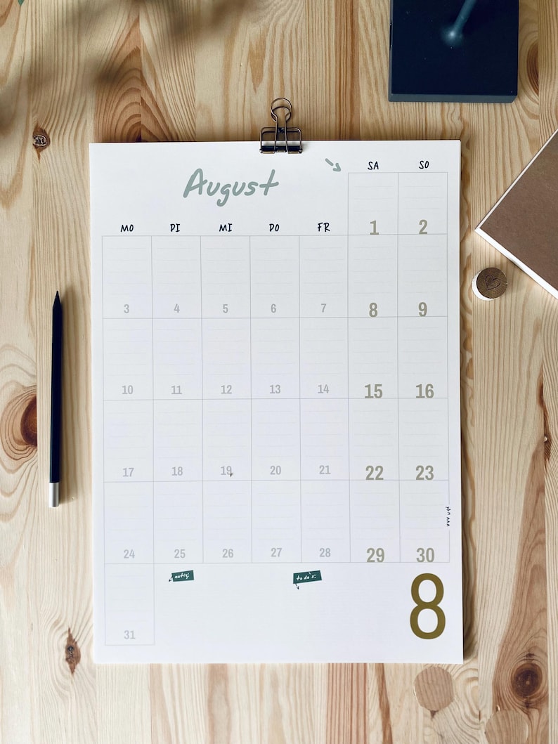 Pode incluir: Um calend&aacute;rio de parede branco para o m&ecirc;s de agosto com um clipe de metal preto no topo. O calend&aacute;rio apresenta um layout de grade com os dias da semana marcados como MO, DI, MI, DO, FR, SA, SO e um grande n&uacute;mero 8 dourado no canto inferior direito.