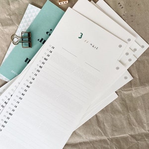 May include: A stack of white paper with a calendar layout, featuring the number "3" and the word "M&auml;rz" in green. The calendar pages have lines for writing and are accompanied by a teal-colored card and a gold paper clip.