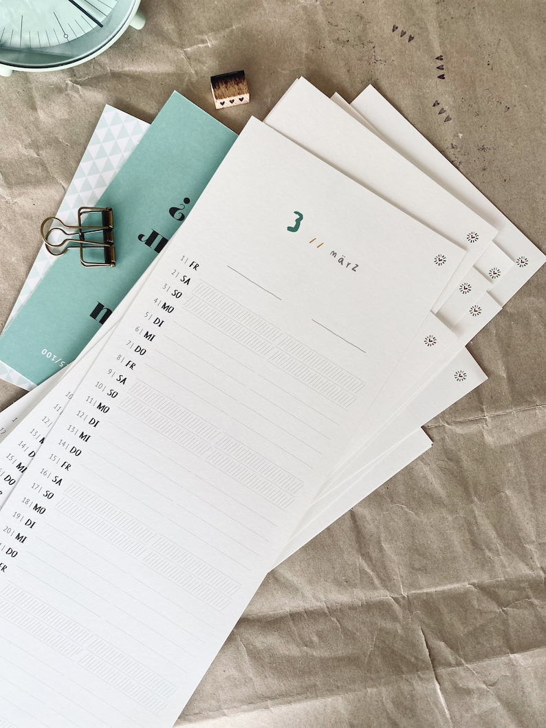 May include: A stack of white paper with a calendar layout, featuring the number "3" and the word "M&auml;rz" in green. The calendar pages have lines for writing and are accompanied by a teal-colored card and a gold paper clip.