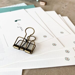 May include: A stack of white stationery with a small green and red floral design. A vintage brass binder clip rests on top of the stationery. A mint green clock and a small wooden block are in the background.