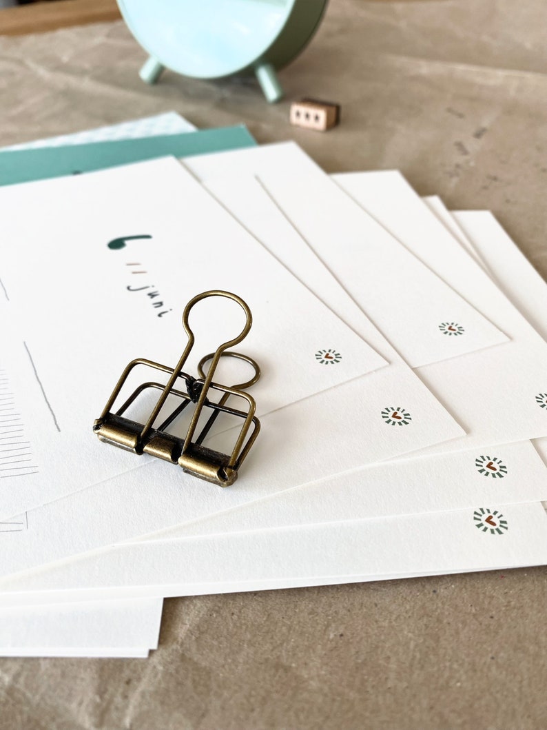 May include: A stack of white stationery with a small green and red floral design. A vintage brass binder clip rests on top of the stationery. A mint green clock and a small wooden block are in the background.