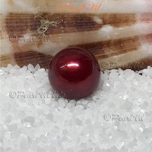 May include: A single, round, red pearl with a high gloss finish. The pearl is sitting on a bed of white, coarse sand.