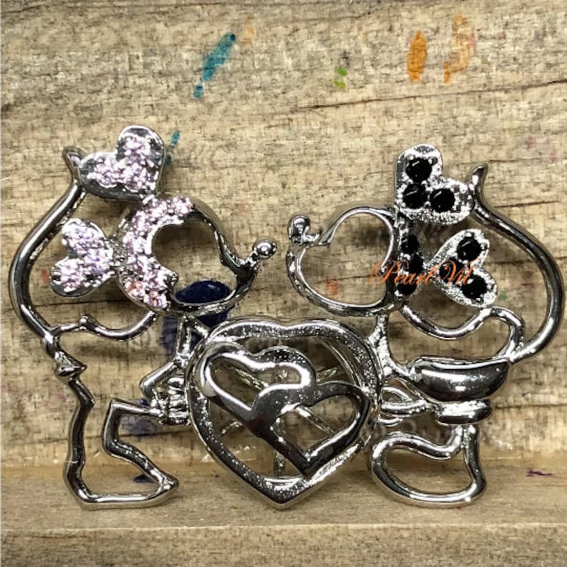 K&ouml;nnte beinhalten: Silberner Anh&auml;nger mit Mickey Mouse und Minnie Mouse Design. Der Anh&auml;nger zeigt zwei ineinander verschlungene Herzen und die Figuren sind mit rosa und schwarzen Strasssteinen verziert.