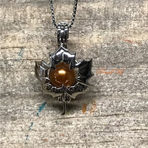 May include: A silver maple leaf charm necklace with a golden pearl inside. The charm is detailed with a textured surface and a delicate stem.
