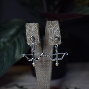 May include: Silver-toned earrings featuring a crossbow design. Each earring has a small ball at the top, a curved bow, and an arrow. The arrowheads are gold-colored. The earrings are displayed on a burlap-textured surface.