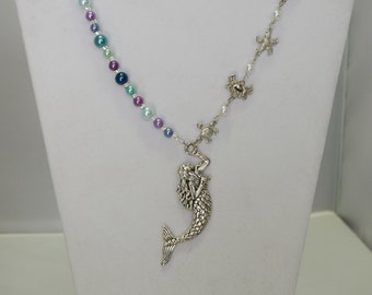 Peut inclure: Un collier pendentif sirène présenté sur un buste blanc. Le collier comporte un rang de perles colorées dans les tons de bleu, violet et vert. Le pendentif est une sirène argentée. Des breloques argentées supplémentaires incluent des fleurs de lys, des étoiles de mer et des tortues.
