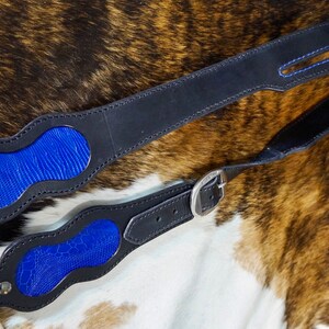 May include: Black leather guitar strap with blue lizard-skin-textured accents and blue stitching. The strap has a silver buckle and rivets. The guitar strap is shown on a cowhide background.