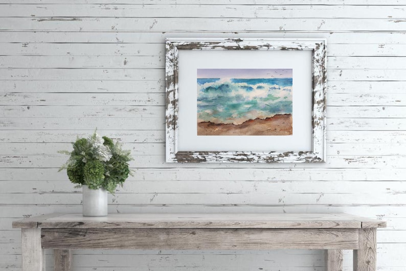 May include: A framed watercolor painting of a seascape with crashing waves, a sandy beach, and a blue sky with a few birds flying overhead.
