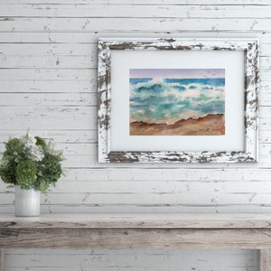 May include: A framed watercolor painting of a seascape with crashing waves, a sandy beach, and a blue sky with a few birds flying overhead.