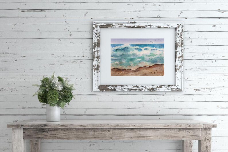 May include: A framed watercolor painting of a seascape with crashing waves, a sandy beach, and a blue sky with a few birds flying overhead.