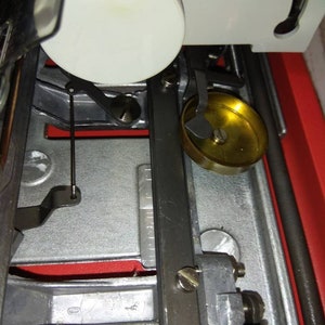 May include: Close-up of a sewing machine with a white plastic spool holder, a black metal arm, and a gold-colored metal bobbin winder.