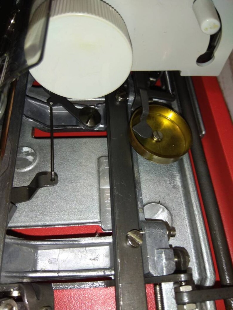 May include: Close-up of a sewing machine with a white plastic spool holder, a black metal arm, and a gold-colored metal bobbin winder.