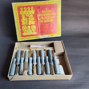 Puede incluir: Un juego de herramientas de talla de madera vintage en una caja de cartón. La tapa de la caja presenta un diseño rojo y amarillo con texto cirílico. En el interior, varios cinceles y gubias con mango gris y marrón están cuidadosamente dispuestos.