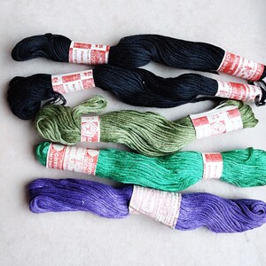 May include: A collection of embroidery floss in various colors, including black, green, and purple. Each skein is wrapped with a paper label featuring red and white text. The floss is arranged on a light-colored surface.