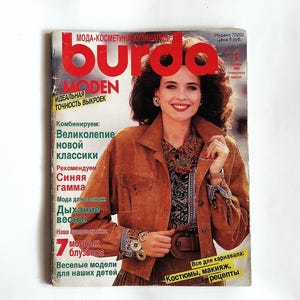 Może przedstawiać: Okładka starego magazynu z kobietą w brązowej zamszowej kurtce. Tytuł magazynu to "Burda Moden" w dużych czerwonych literach. Okładka zawiera tekst w języku rosyjskim, reklamujący modę i urodę.
