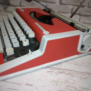 May include: A red and white vintage typewriter with a white keyboard and black keys. The typewriter has a carriage return lever and a paper roller.