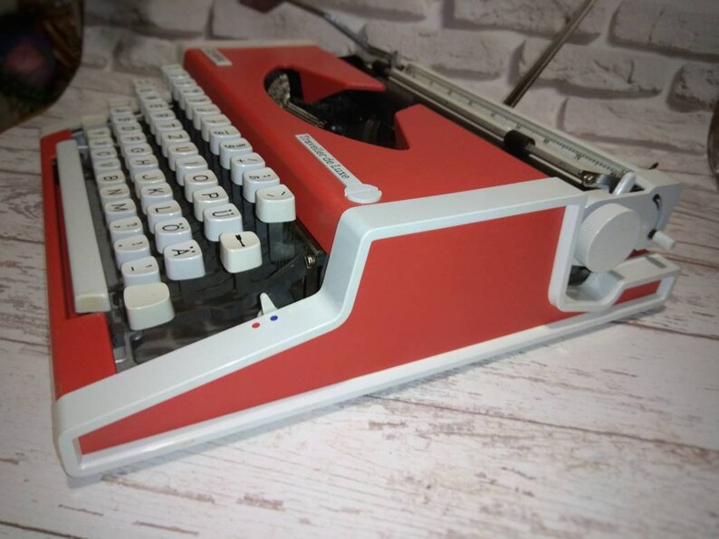 May include: A red and white vintage typewriter with a white keyboard and black keys. The typewriter has a carriage return lever and a paper roller.