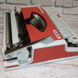 May include: A red and white vintage Olympia typewriter with white keys and a silver carriage return lever. The typewriter has a red body and a white base.