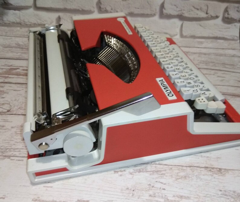 May include: A red and white vintage Olympia typewriter with white keys and a silver carriage return lever. The typewriter has a red body and a white base.