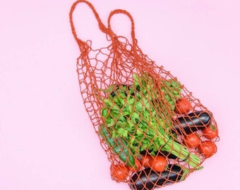 May include: A brown, open-weave net bag filled with fresh produce, including tomatoes, eggplants, and parsley. The bag has two handles and is set against a light pink background.