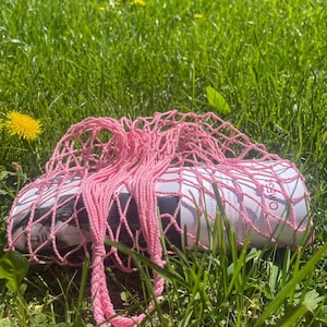 May include: Pink string mesh bag with a white and black magazine inside. The bag is lying on green grass with yellow flowers.