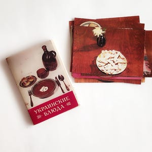May include: A vintage cookbook with a red and white cover, titled in Russian. The book is next to a stack of wooden placemats, each with a photo of food. One placemat shows a plate of pastries and a small vase with a flower.