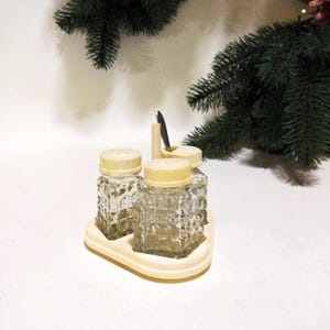 Puede incluir: Un juego de tres saleros y pimenteros de vidrio transparente con tapas blancas, que se encuentran sobre una base de plástico blanco con una pequeña cuchara.