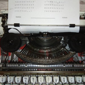 May include: A white vintage typewriter with a red and black design. The typewriter has a sheet of paper inserted and the keys are visible. The keys are labeled with numbers, symbols, and letters. The text on the paper reads: "123456789+3", "qwertyuiop", "asdfghjkl", "zxcvbnm", "1- & ( ) - _ / ", "QWERTYUIOPU", "ASDFGHJKL", "ZXCVBNM", "1".