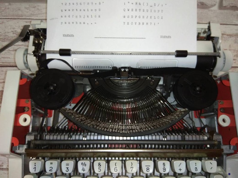 May include: A white vintage typewriter with a red and black design. The typewriter has a sheet of paper inserted and the keys are visible. The keys are labeled with numbers, symbols, and letters. The text on the paper reads: "123456789+3", "qwertyuiop", "asdfghjkl", "zxcvbnm", "1- & ( ) - _ / ", "QWERTYUIOPU", "ASDFGHJKL", "ZXCVBNM", "1".