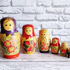 May include: A set of five traditional Russian nesting dolls, or matryoshka dolls. The dolls are painted in bright colors with floral patterns and have smiling faces. The largest doll is red, white, and blue, and the smallest doll is yellow and red.
