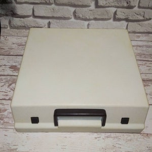 May include: A white vintage typewriter case with a brown handle and two black buttons on the front.