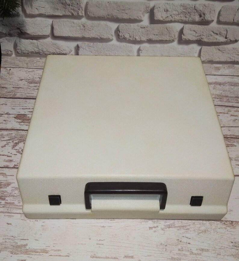 May include: A white vintage typewriter case with a brown handle and two black buttons on the front.