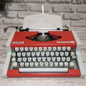 May include: A red and white vintage Olympia Traveller de Luxe typewriter with a white paper insert. The typewriter has a black roller and a black and white keyboard with the letters QWERTYUIOPASDFGHJKL&Ouml;&Auml;YXCVBNM and numbers 1234567890.