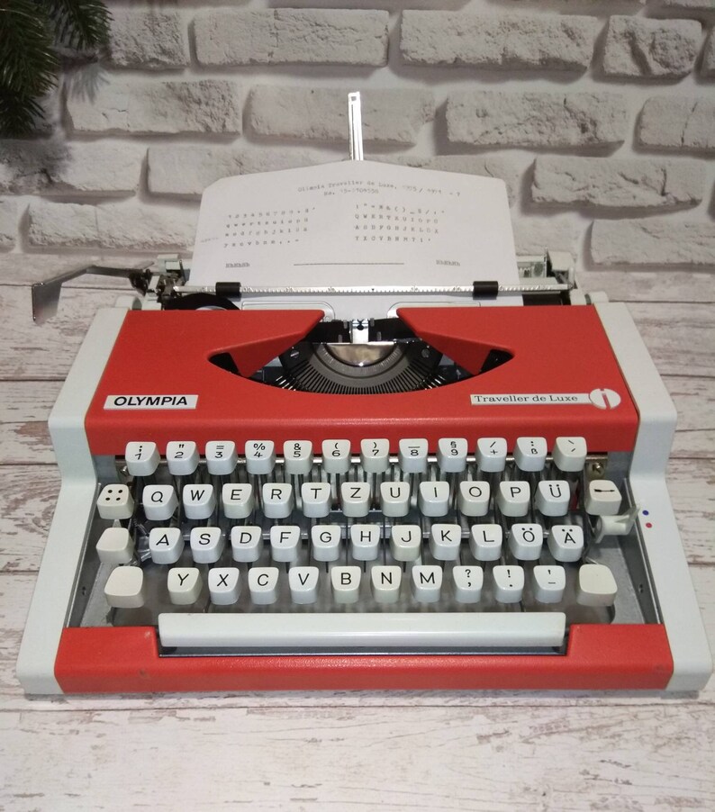May include: A red and white vintage Olympia Traveller de Luxe typewriter with a white paper insert. The typewriter has a black roller and a black and white keyboard with the letters QWERTYUIOPASDFGHJKL&Ouml;&Auml;YXCVBNM and numbers 1234567890.