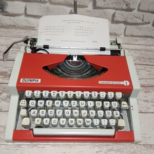May include: A red and white Olympia Traveller de Luxe typewriter with a white paper insert. The typewriter has a QWERTY keyboard with German characters, including "&Ouml;", "&Auml;", and "&Uuml;".