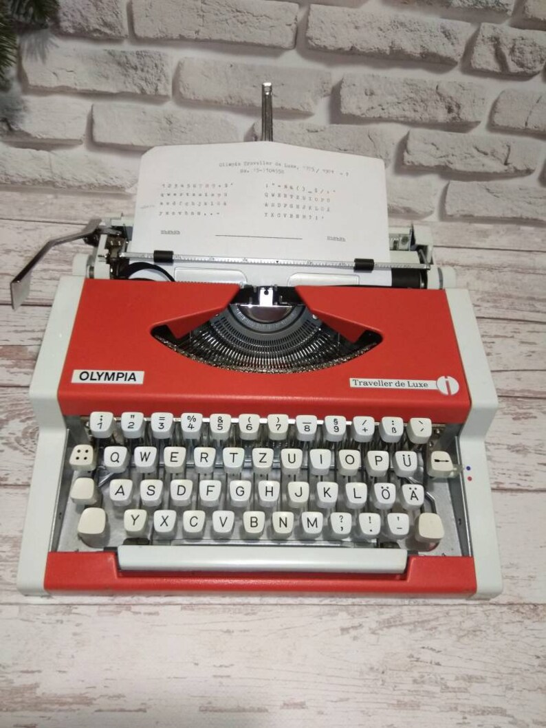 May include: A red and white Olympia Traveller de Luxe typewriter with a white paper insert. The typewriter has a QWERTY keyboard with German characters, including "&Ouml;", "&Auml;", and "&Uuml;".