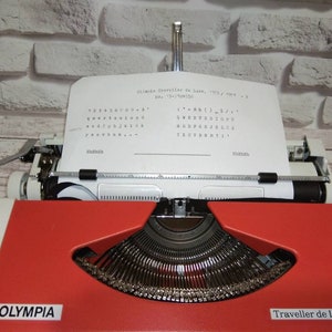 May include: A red and white Olympia Traveller de Luxe typewriter with a sheet of paper inserted. The paper has the text "Olympia Traveller de Luxe, "705" / "994" - 1" and a keyboard layout printed on it.