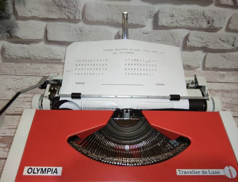 May include: A red and white Olympia Traveller de Luxe typewriter with a sheet of paper inserted. The paper has the text "Olympia Traveller de Luxe, "705" / "994" - 1" and a keyboard layout printed on it.