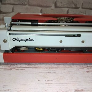 May include: A red and white vintage Olympia typewriter. The typewriter has a metal carriage and a black label with the word "Olympia" printed on it.