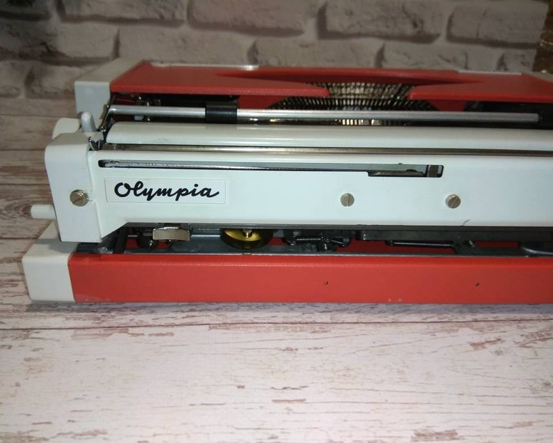 May include: A red and white vintage Olympia typewriter. The typewriter has a metal carriage and a black label with the word "Olympia" printed on it.