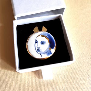 May include: A circular brooch featuring a blue and white painted portrait of a face, set in a gold-toned metal frame with two small gold-toned tabs at the top, presented inside a white box with a black velvet insert.
