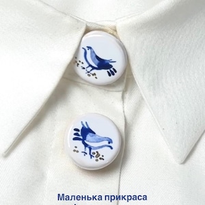 May include: Two round white buttons with blue bird illustrations attached to a white collared shirt. The text "Маленька прикраса змінює враження" is printed below the buttons. The buttons feature a glossy finish and detailed blue avian motifs.