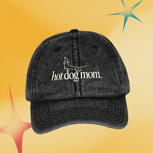 Puede incluir: Gorra de b&eacute;isbol negra con el texto "hot dog mom" en escritura blanca. La gorra presenta una ilustraci&oacute;n de perro salchicha encima del texto. El sombrero est&aacute; hecho de una tela lavada y texturizada y tiene una visera curva.
