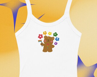 Puede incluir: Una camiseta de tirantes blanca con un gráfico central de un oso marrón que sostiene una pequeña bandera arcoíris. El oso está rodeado por un semicírculo de flores coloridas en rojo, naranja, amarillo, verde y azul. El oso tiene mejillas rosadas.