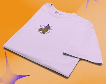 Puede incluir: Una camiseta de manga corta de color morado claro con un pequeño gráfico bordado en el pecho izquierdo. El gráfico muestra un perro salchicha marrón con un sombrero de bruja azul oscuro, situado en el centro de un diseño de telaraña morada.