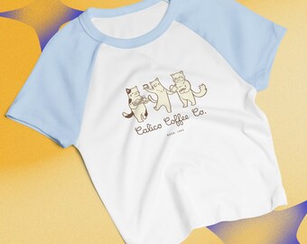 Puede incluir: Camiseta raglán blanca y azul claro con un gráfico de tres gatos de dibujos animados sosteniendo tazas de café y una bandeja. El texto "Calico Coffee Co. Estd. 1994" está impreso debajo. La camiseta tiene mangas azules claras y cuello azul claro.