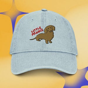 Puede incluir: Una gorra de béisbol de mezclilla azul claro con un perro salchicha marrón bordado. Sobre el perro, el texto "LITTLE MENACE" está bordado en hilo rojo. La gorra tiene una visera curva, un botón en la parte superior y ojales de ventilación.