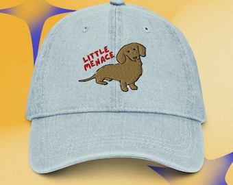 Puede incluir: Una gorra de béisbol de mezclilla azul claro con un perro salchicha marrón bordado. Sobre el perro, el texto "LITTLE MENACE" está bordado en hilo rojo. La gorra tiene una visera curva, un botón en la parte superior y ojales de ventilación.