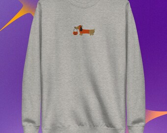 Puede incluir: Una sudadera gris jaspeado de cuello redondo con un pequeño gráfico centrado de un perro salchicha marrón con un sombrero azul y un pequeño cubo de dulces naranja en forma de calabaza. La prenda de manga larga tiene cuello y puños acanalados.