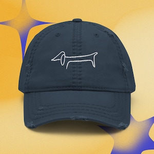Puede incluir: Una gorra de béisbol azul marino desgastada con un gráfico bordado en hilo blanco de un perro salchicha centrado en el panel frontal. La gorra tiene una visera curva con bordes deshilachados y una corona estructurada con botón superior.